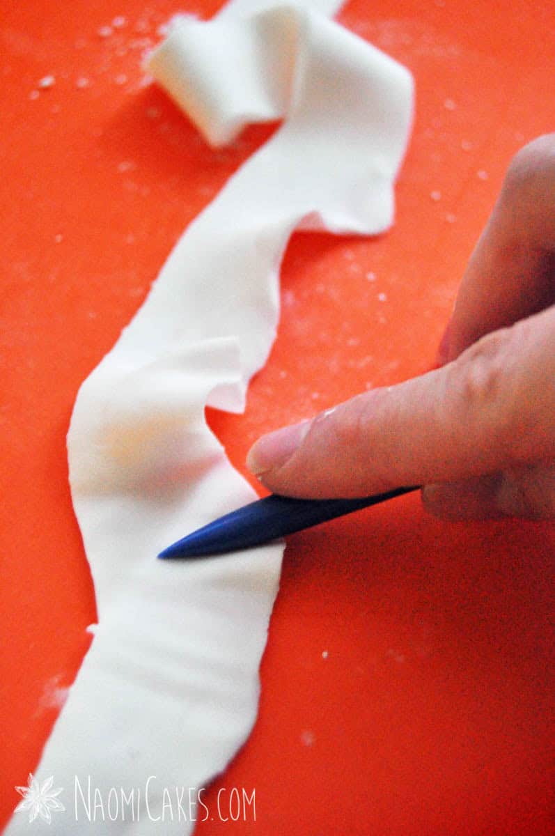 a hand making a ruffled strip of gum paste/fondant on an orange mat