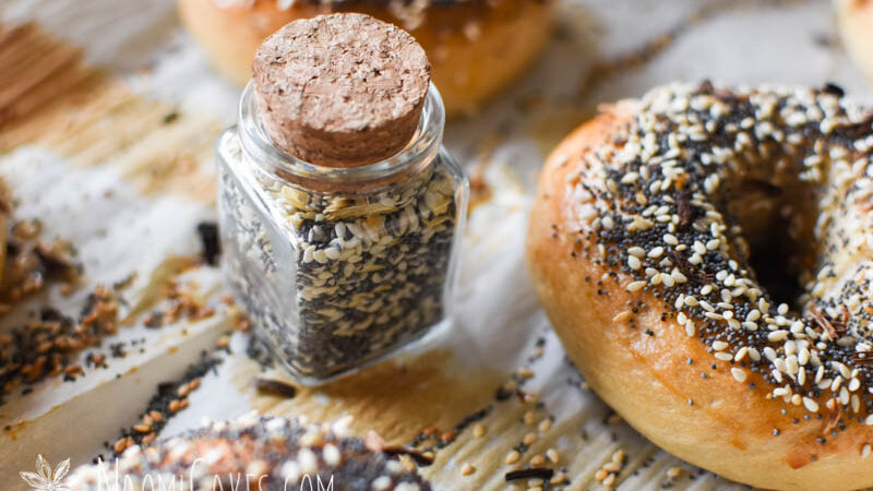 close up closed cork lid jar of everything bagel seasoning on a pan with fresh baked everything bagels