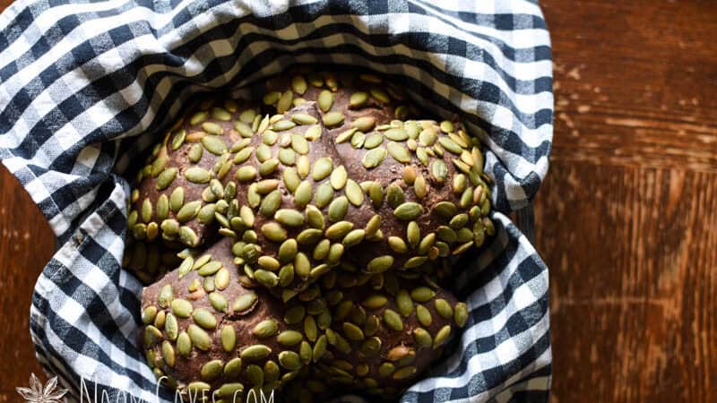 baked dark rye pumpkinseed covered rolls in a bowl with a checked cloth